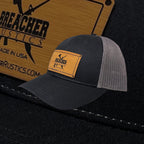 Breacher Rustics black cotton trucker hat with grey mesh back and bamboo leather patch. Structured, low-profile with snapback closure.