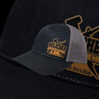 Breacher Rustics black trucker hat with a grey mesh back and a wooden patch featuring crossed tools and the words "Breacher Rustics Made in USA".