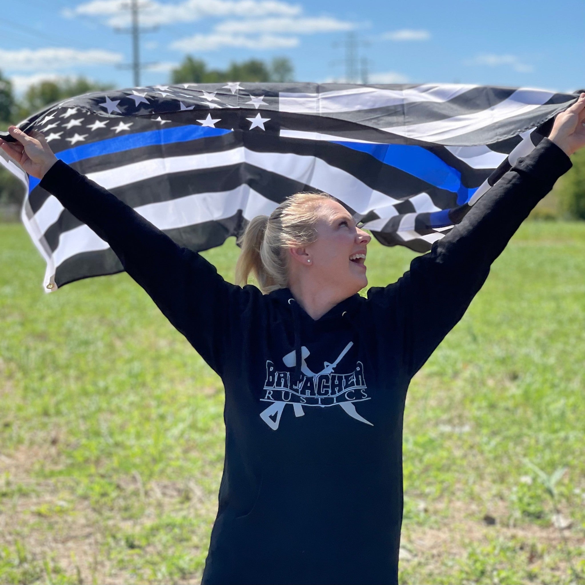 Breacher Rustics black cotton blend pullover hoodie with muff pocket and logo, worn by a woman holding a thin blue line flag. breacher rustics patriotic hoodie american flag apparel first responder clothing soft durable hoodie usa made